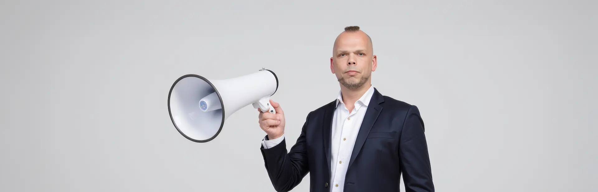 Mann in Anzug hält ein Megafon in der Hand und schaut direkt in die Kamera.